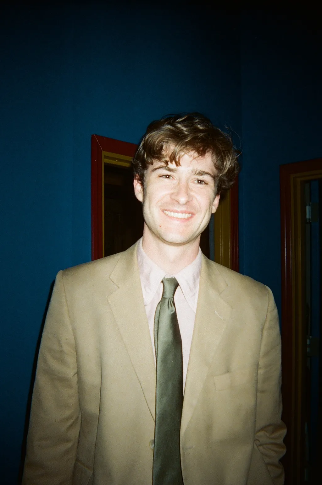 Andrew Hardigg smiling in a tan suit and green tie in front of a deep blue wall
