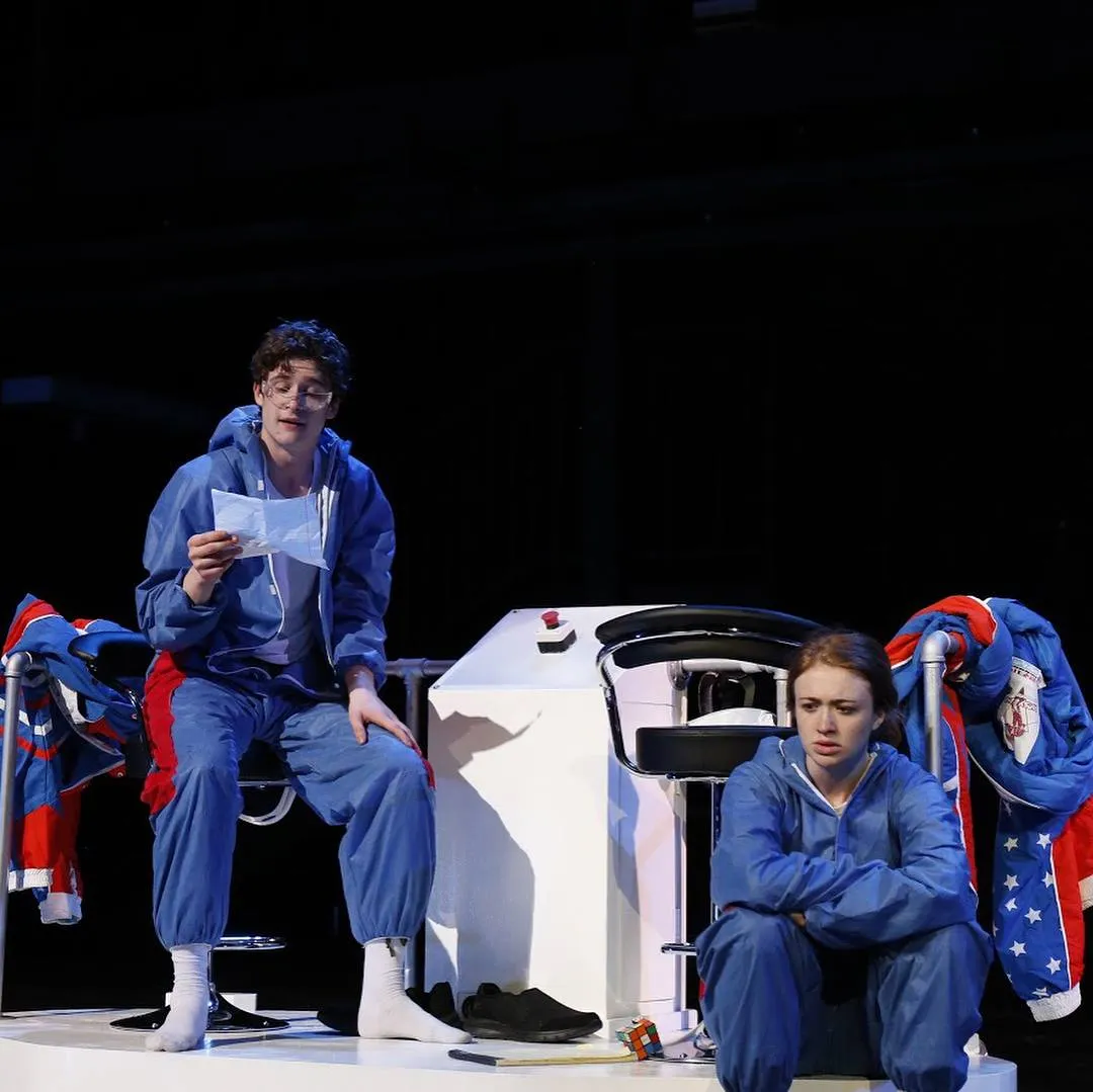 Andrew Hardigg in a blue jumpsuit reading from a page on a darkened stage