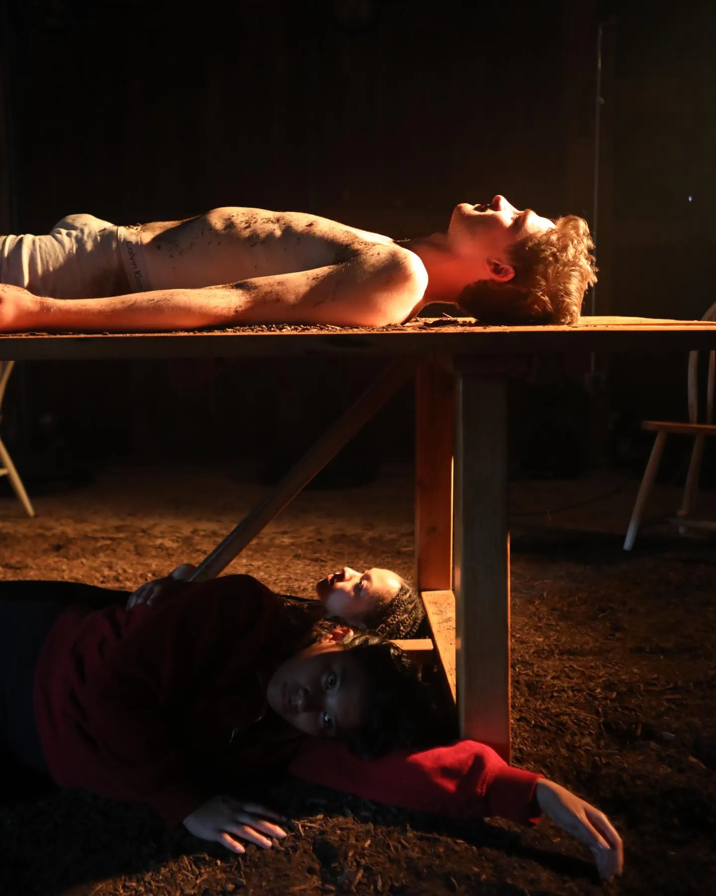 Andrew Hardigg lying across a wooden table under warm light, two actors on the ground below