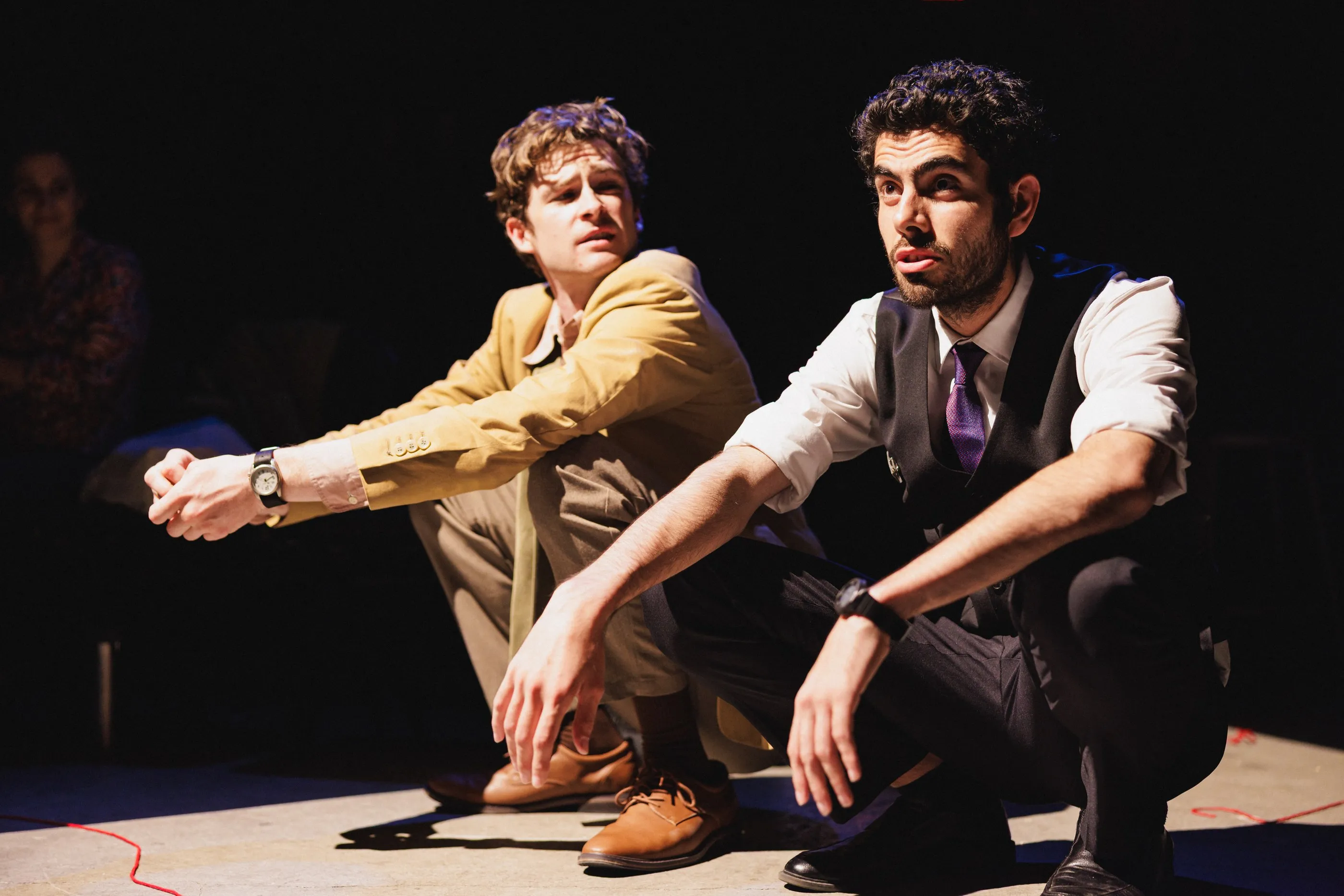 Andrew Hardigg on stage in a tan jacket, crouched beside another actor under warm light